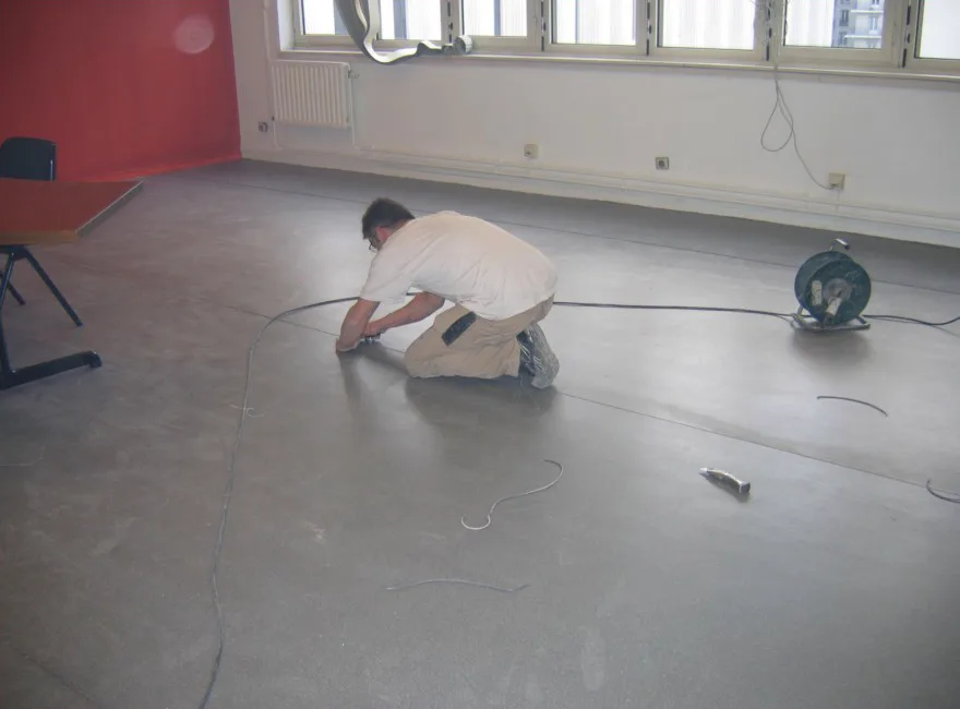 Un poseur à genoux déroule et ajuste un sol PVC en lés sur un plateau de bureaux vide
