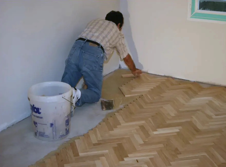 Un poseur assemble un parquet en point de Hongrie, au pied d'un mur, à côté d'un seau de colle