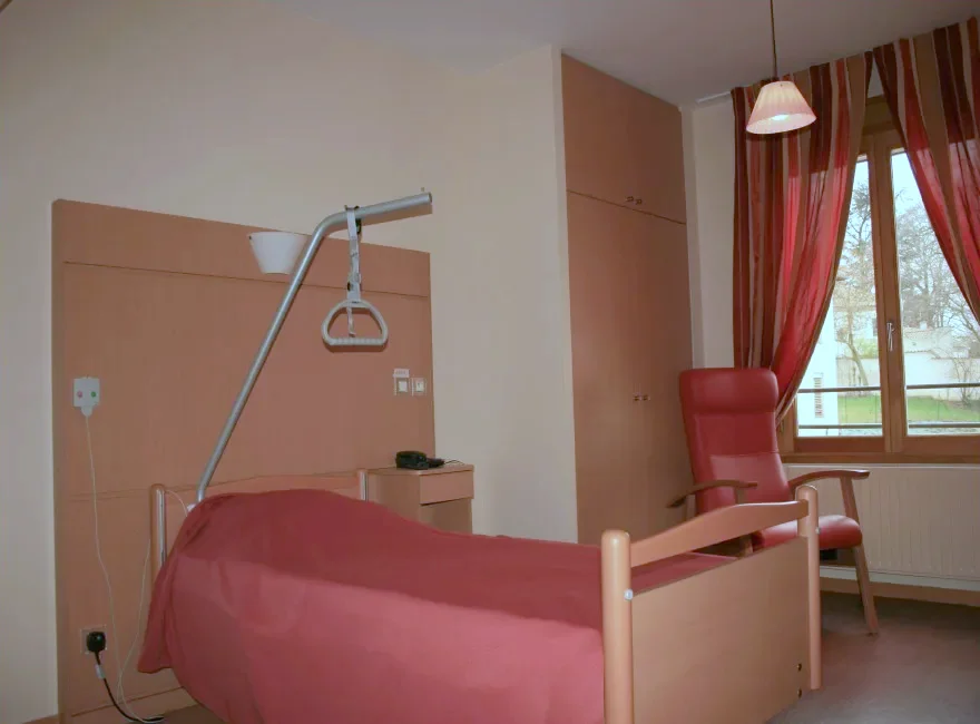 Chambre médicalisée d'une résidence pour personnes âgées : lit, fauteuil rouge, sol souple