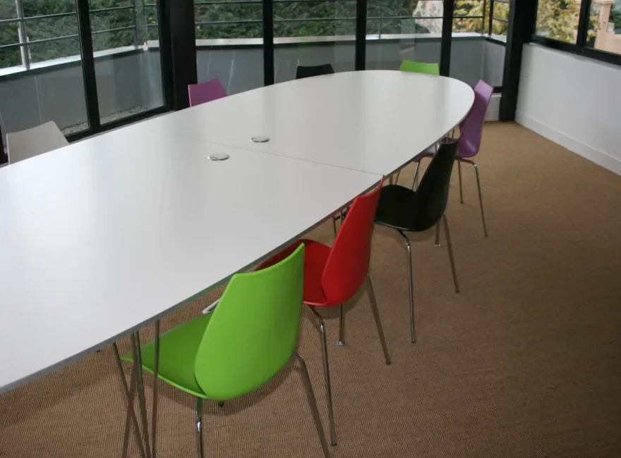 Salle de réunion de bureaux : grande table blanche, chaises colorées, sol textile